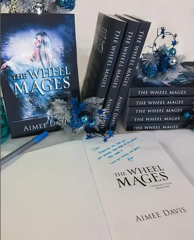 Pile of books of The Wheel Mages (black cover with white woman in white dress surrounded by water) with decorative silver and blue ornaments. One book is open on the table with a pen nearby.
Copyright: Aimee Davis
Source: Instagram @writingwaimee 