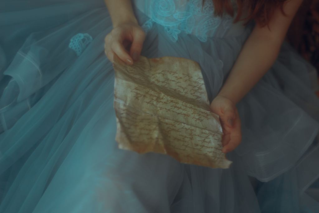 White hands hold a piece of piece of parchment against a lap draped in a blue dress.
Source: Unplash.com 